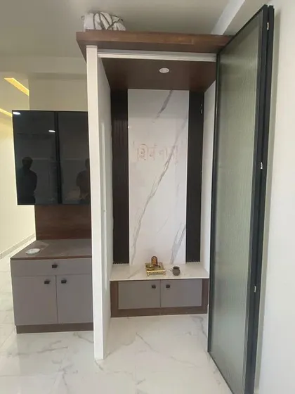A compact pooja unit integrated seamlessly into the living area. This design features a marble-finish back panel and is enclosed by a modern, foldable glass door.