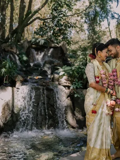 A breathtaking shot of the couple at Miraya Greens. The natural waterfall provides a stunning backdrop for their traditional South Indian wedding attire, creating a moment of pure tranquility.