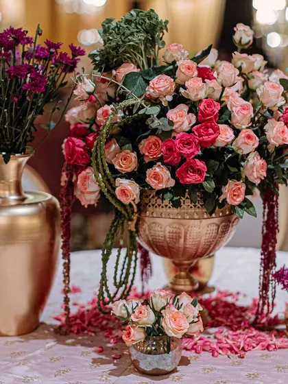 A table centerpiece with an arrangement of pink and red roses in a golden urn, surrounded by scattered petals. This detail shot highlights the romantic and delicate floral work of the event.