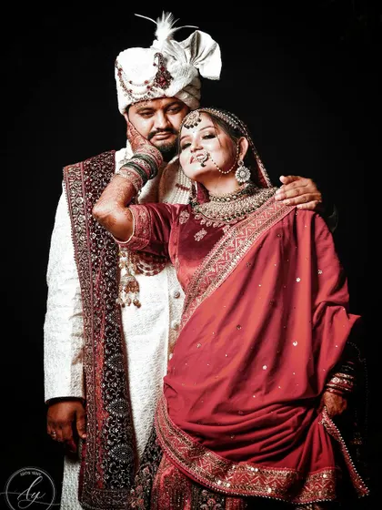A stylish and confident wedding portrait. The bride playfully holds her groom's face, showcasing her intricate henna and jewelry, while he looks on with affection. The dark background makes their vibrant attire pop.