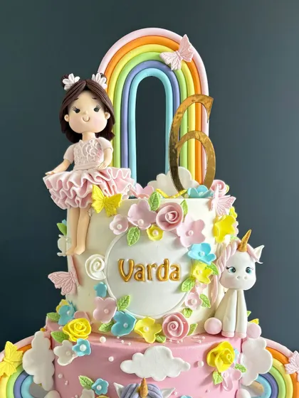 Unleash the magic! This two-tiered unicorn birthday cake is a pastel dream, featuring a sweet doll, a unicorn, and a vibrant rainbow for Varda's 6th birthday.