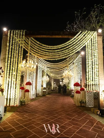 A grand, traditional Indian wedding entrance featuring swags of fresh white flowers, crystal chandeliers, and a rich, patterned carpet. This design creates a sense of royal arrival and celebrates our rich cultural heritage.