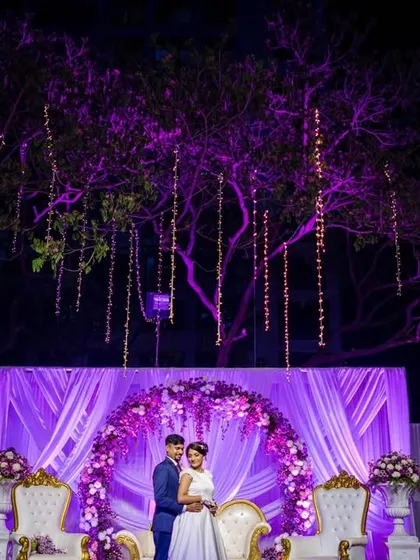 A grand reception stage with the couple framed by an arch of purple flowers and hanging lights. This portrait showcases the beautiful decor and scale of the event.