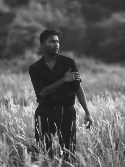 A strong black and white portrait of a man in a dark shirt standing in a field. His gaze is directed away from the camera, creating a candid and masculine portrait with a natural feel.