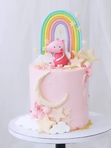 A sweet and simple Peppa Pig cake. This pink design features a fondant Peppa figure under a pastel rainbow, surrounded by a crescent moon and stars.