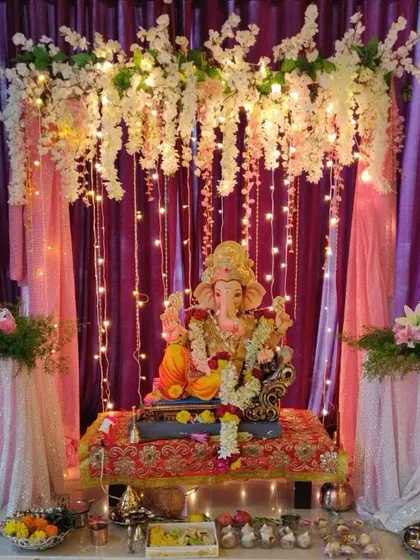 A dreamy Ganpati mandap created with a canopy of white flowers and cascading fairy lights against a purple curtain. The sequined fabric on the side tables adds a soft, celebratory shimmer.