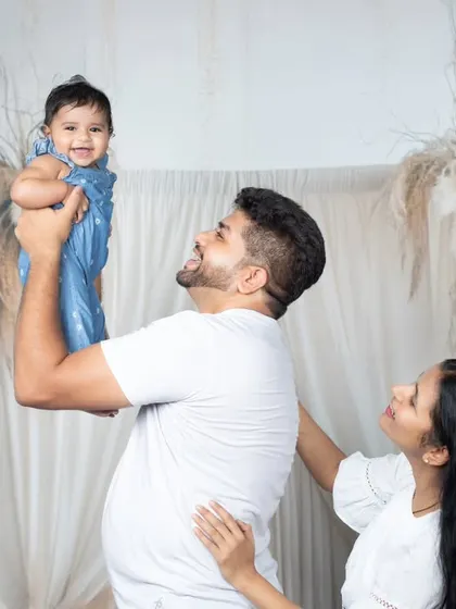A father joyfully lifts his baby daughter high in the air as the mother watches with a smile.