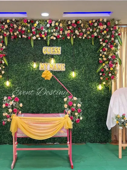 This setup includes a decorated cradle placed before a green wall backdrop. The "Guess My Name" sign and hanging bulbs add a modern and playful touch to the traditional ceremony.