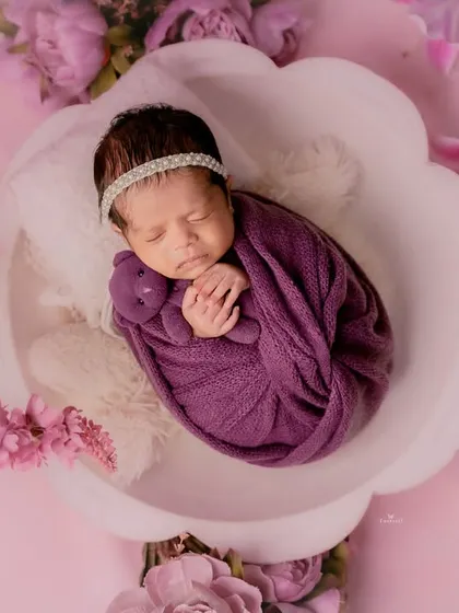 A beautiful floral-themed newborn portrait. The baby is swaddled in purple and rests in a flower-shaped bowl, surrounded by soft pink lilies.