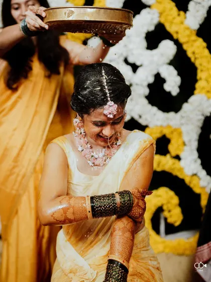 The bride smiles as water is poured over her during the Haldi ceremony, her floral jewelry still intact.