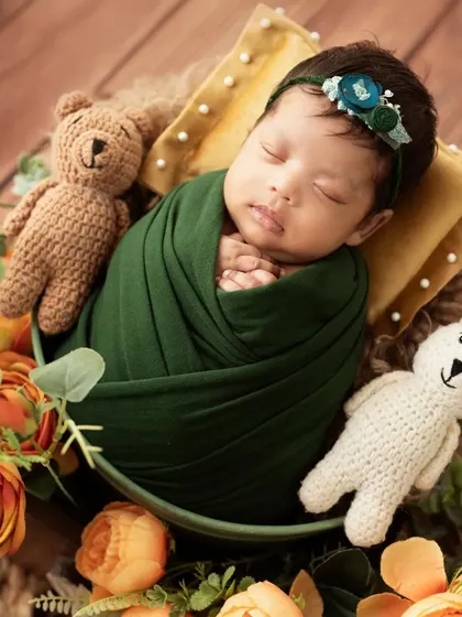 A sleeping newborn wrapped in green, surrounded by orange flowers and holding two tiny teddy bears.