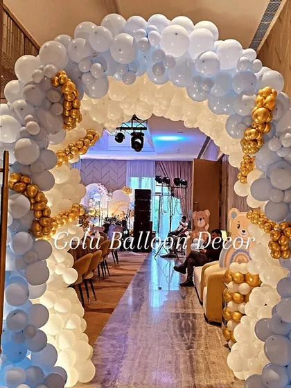A magnificent balloon arch in white, pastel blue, and gold creates a grand entrance for a teddy bear-themed first birthday party.