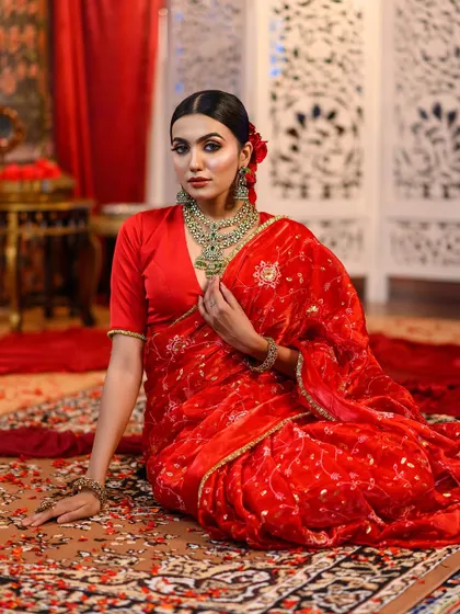 A luxurious portrait featuring a red velvet saree. The model is seated on an ornate carpet, creating a scene of royal opulence.