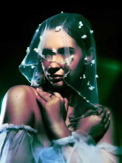 A soft, intimate portrait where the model clutches her shoulders, her face obscured by the pearl veil. The image is both vulnerable and enigmatic.