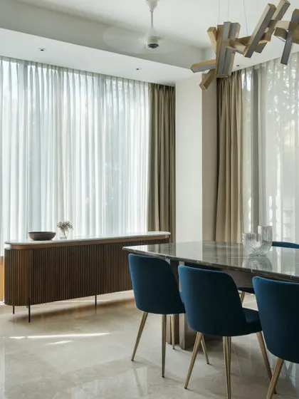 The dining area is designed as a bright, airy space for gathering. A sculptural wooden light fixture hangs above the marble-topped table, while sheer curtains diffuse the sunlight, creating a soft glow. The material palette of wood, stone, and rich blue upholstery adds layers of texture and warmth.