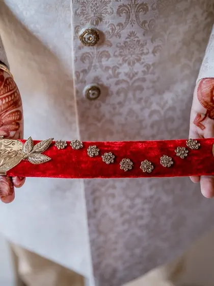A close-up on the groom's hands, showcasing his intricate henna and the detailed velvet scabbard of his sword, key elements of his wedding attire.
