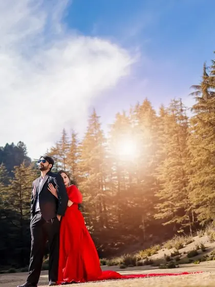 A romantic portrait of a couple in a pine forest, with the woman in a striking red gown leaning against her partner as the sun creates a beautiful flare.