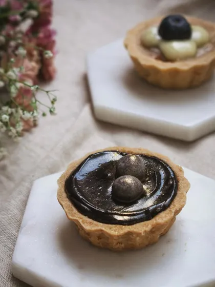 A pair of gourmet tarts, one chocolate and one fruit, on minimalist marble coasters. The soft lighting and simple styling emphasize the fine details and artistry of the pastries.