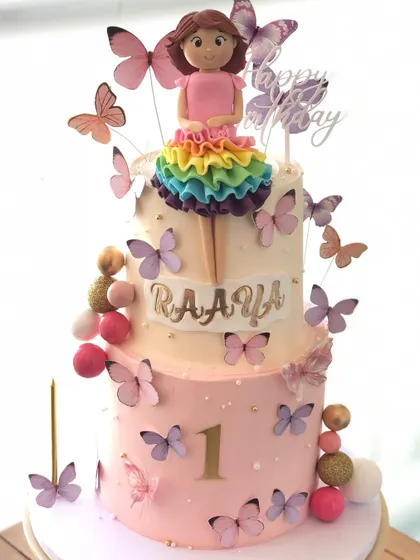 A two-tiered butterfly-themed cake for a first birthday, with a rainbow-dress doll topper and a flutter of edible butterflies.