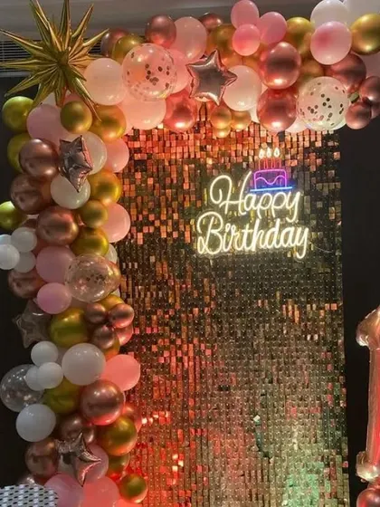 A birthday party with a rose gold shimmer wall, a 'Happy Birthday' neon sign, and a balloon garland.