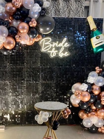 A chic bachelorette party backdrop. The decor features a black sequin wall, a "Bride to be" neon sign, and a balloon arrangement in black, white, and rose gold.