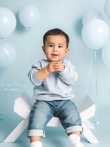 A handsome little man in jeans and a sweater, sitting confidently for his milestone portrait. The simple blue background makes his happy expression stand out.