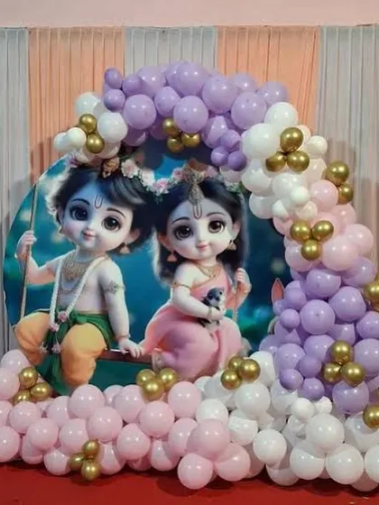 A beautiful baby shower backdrop featuring adorable illustrations of baby Krishna and Radha. The scene is framed by a pastel balloon arch in pink, purple, and white, with small gold balloon accents.