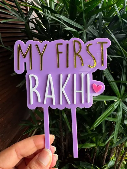Holding our "My First Rakhi" topper to show its size and vibrant colors. The gold mirror layer adds a touch of festive shine.