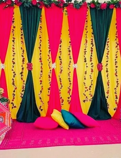 A colorful stage setup with a backdrop of yellow and green drapes tied back with floral arrangements. The floor is covered with a bright pink carpet and colorful cushions.