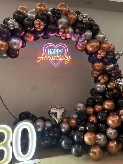 I design modern and stylish decorations for milestone celebrations like a 30th anniversary. This setup includes a sophisticated balloon arch in black, silver, and bronze chrome, a 'Happy Anniversary' neon sign, and light-up '30' numbers.