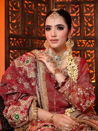A detailed portrait of a model in a rich red bridal outfit. The shot emphasizes the heavy earrings, maang tikka, and the intricate handiwork on the dupatta.