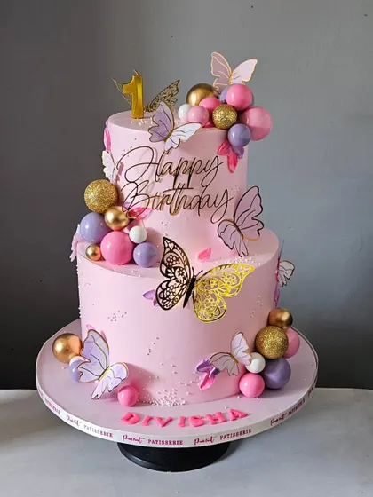 One year of pure joy, wrapped in pink and butterflies! This two-tier first birthday cake is adorned with delicate butterflies and decorative spheres in shades of pink, purple, and gold.