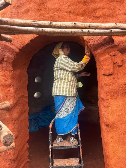 A mason stands on a ladder, carefully applying a finish to the arched entrance of a mud structure. Her focus and craftsmanship are evident in every detail.