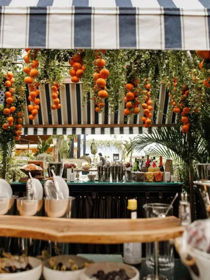 A close-up of the bar at a garden soiree. The hanging oranges and lush greenery create a vibrant and fresh canopy over the drink station, tying into the event's citrus theme.