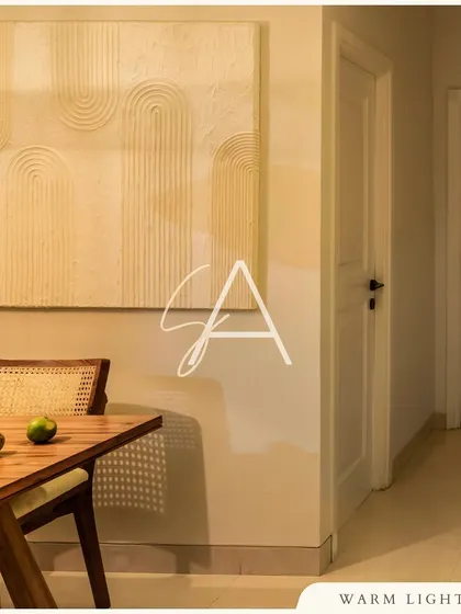 The dining area features a textured art piece that adds depth and interest to the minimalist space. The warm lighting and natural wood of the cane chair create a simple yet elegant setting for meals.