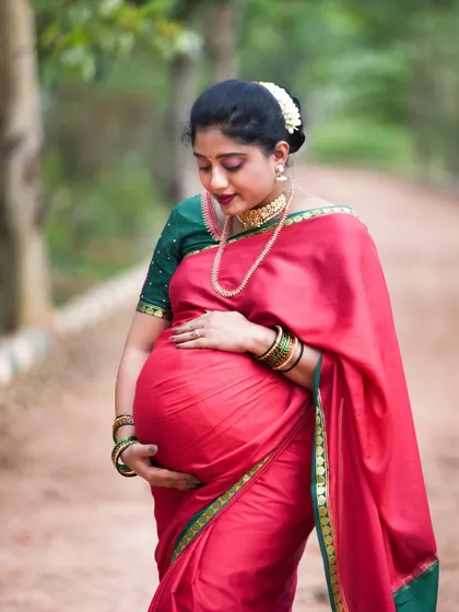 An elegant outdoor portrait of a mom-to-be in a traditional red saree, capturing a moment of quiet reflection.