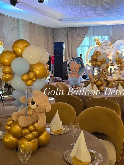 An elegant table centerpiece for a teddy bear-themed party, featuring a teddy bear holding a bouquet of gold, white, and pastel blue balloons.