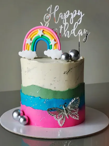 A colorful rainbow-themed birthday cake. The design features a multi-colored buttercream base, a bright rainbow topper, and a silver butterfly for a touch of sparkle.