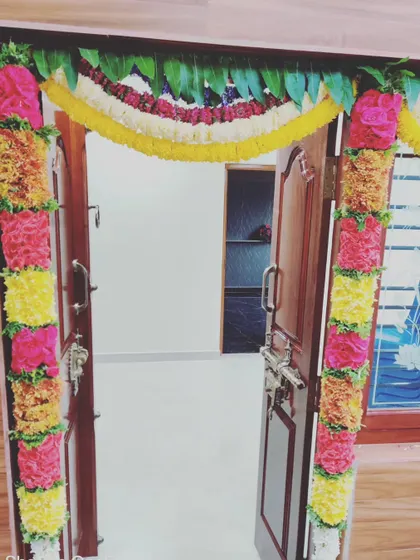 A beautifully decorated doorway for a housewarming event. The door frame is lined with colorful flower arrangements and a traditional mango leaf toran at the top, signifying prosperity and good fortune.