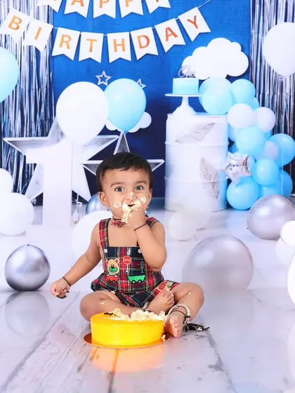 A happy baby boy enjoys his yellow birthday cake during a cake smash session. The blue, silver, and white balloon backdrop creates a festive and celebratory mood.