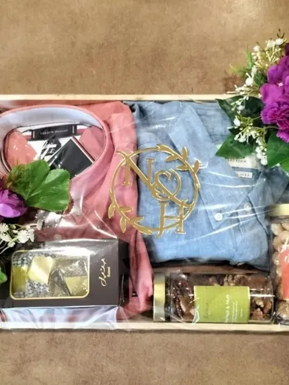 A custom gift box designed to hold multiple apparel items for the groom. This arrangement includes two shirts, gourmet treats, and a personalized monogram, all accented with rich purple floral bouquets.