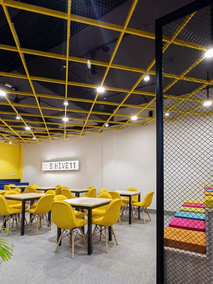 The yellow grid ceiling is a dominant feature in this cafeteria space, adding an industrial yet playful element. It defines the area and complements the bright yellow seating, creating a sunny and inviting atmosphere.