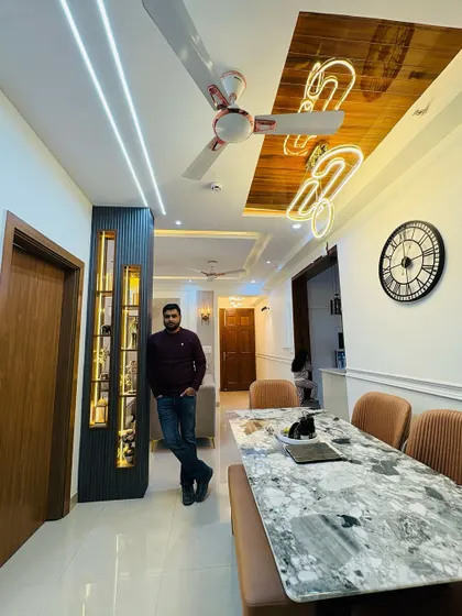 A view of the dining area in a 3BHK project. The space features a marble-top dining table, a custom partition with display shelves, and a unique neon-style light fixture on the wooden ceiling panel.