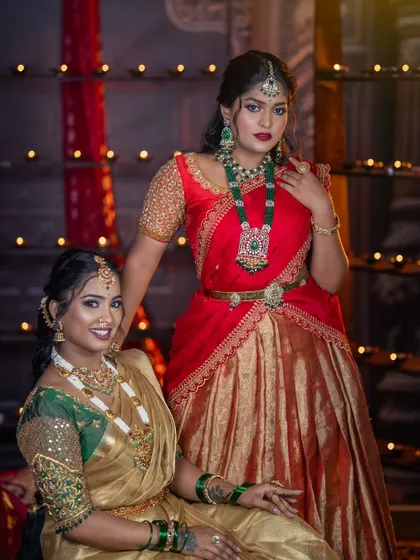 A full shot of the two friends, showcasing their coordinated yet individual styles. One is in a red lehenga with a modern emerald set, and the other is in a gold saree with traditional jewellery.