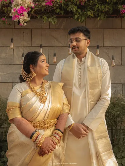 A portrait of a couple in matching white and gold traditional wear. Their comfortable smiles and shared look speak of a deep and happy connection.