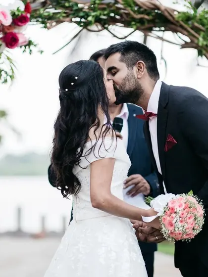A romantic kiss seals the vows under a rustic vine and floral arch. This intimate setup by the water was perfect for a beautiful and personal Catholic wedding ceremony.