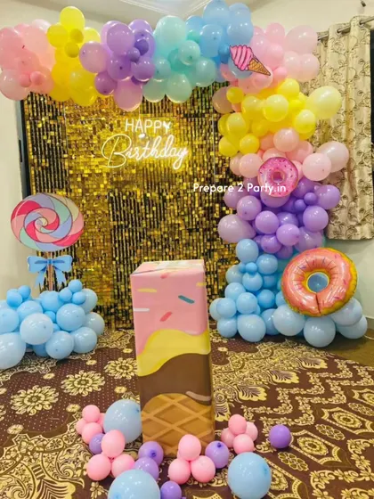 A delicious-looking Candyland theme decoration. This sweet setup features a gold sequin wall, a pastel balloon garland, and large props like a lollipop, donut, and an ice cream cone cake stand.