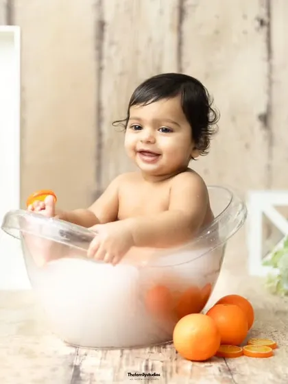 A baby girl plays with an orange slice in the tub, adding a fun, interactive element to the photoshoot.