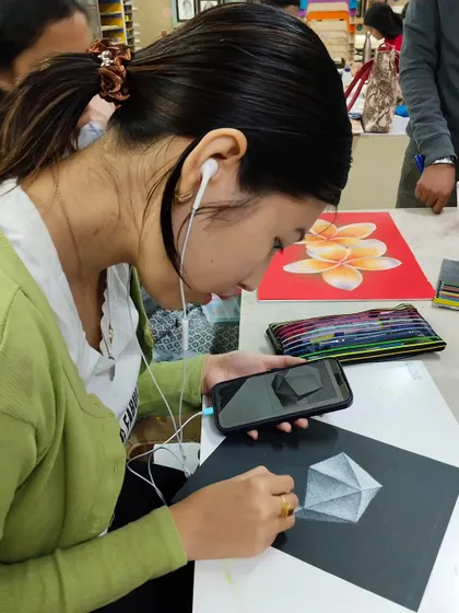 A student using her phone for reference while working on a detailed drawing with pastels, blending modern tools with traditional techniques.