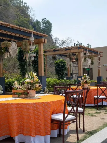 An outdoor dining area with a rustic and bohemian feel. The tables are covered in bright orange cloths, and the wooden pergolas are decorated with hanging macrame and floral elements.